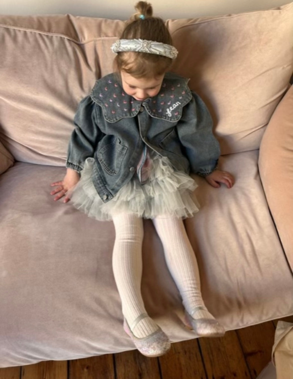 Child wearing a denim jacket and tulle skirt sitting on a pink couch.