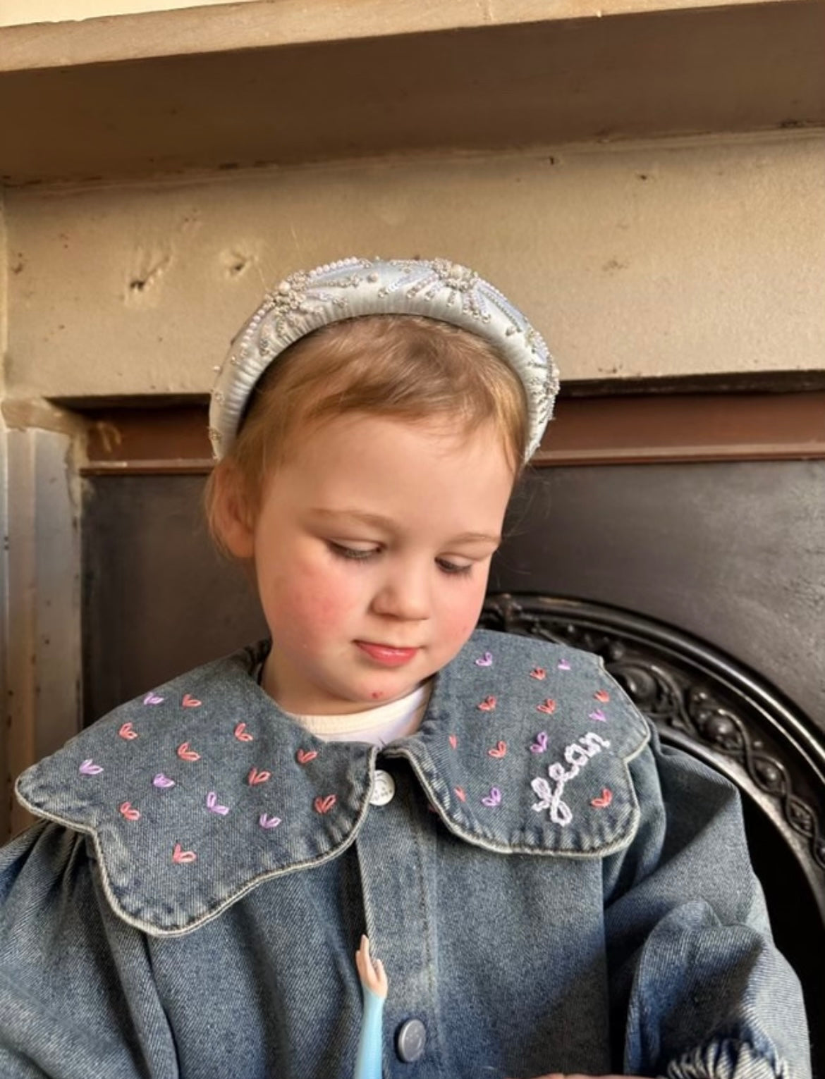 Child wearing a denim jacket with embroidered collar and bonnet, sitting in front of a fireplace.