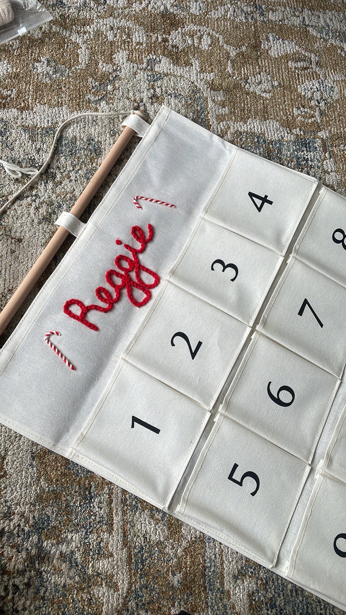 Two personalized Christmas advent calendars laid out on a patterned rug over a wood floor. One calendar is white with the name 'George' in red script and the other is white with the name 'Ruby' in red script, both with numbered pockets and wooden dowels