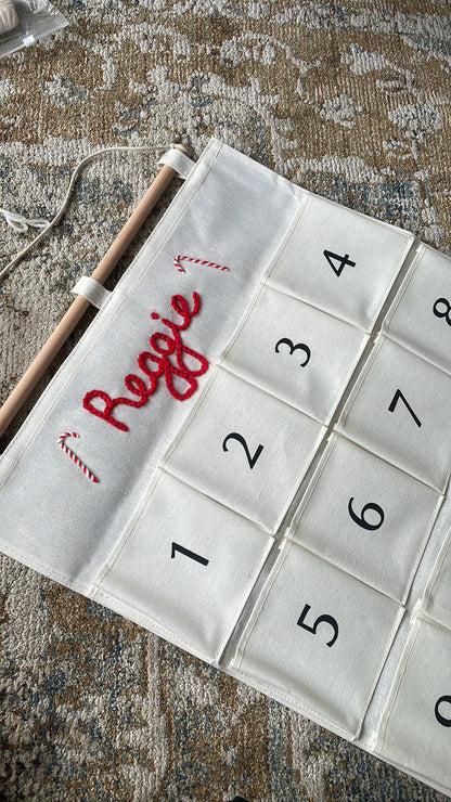 Two personalized Christmas advent calendars laid out on a patterned rug over a wood floor. One calendar is white with the name 'George' in red script and the other is white with the name 'Ruby' in red script, both with numbered pockets and wooden dowels