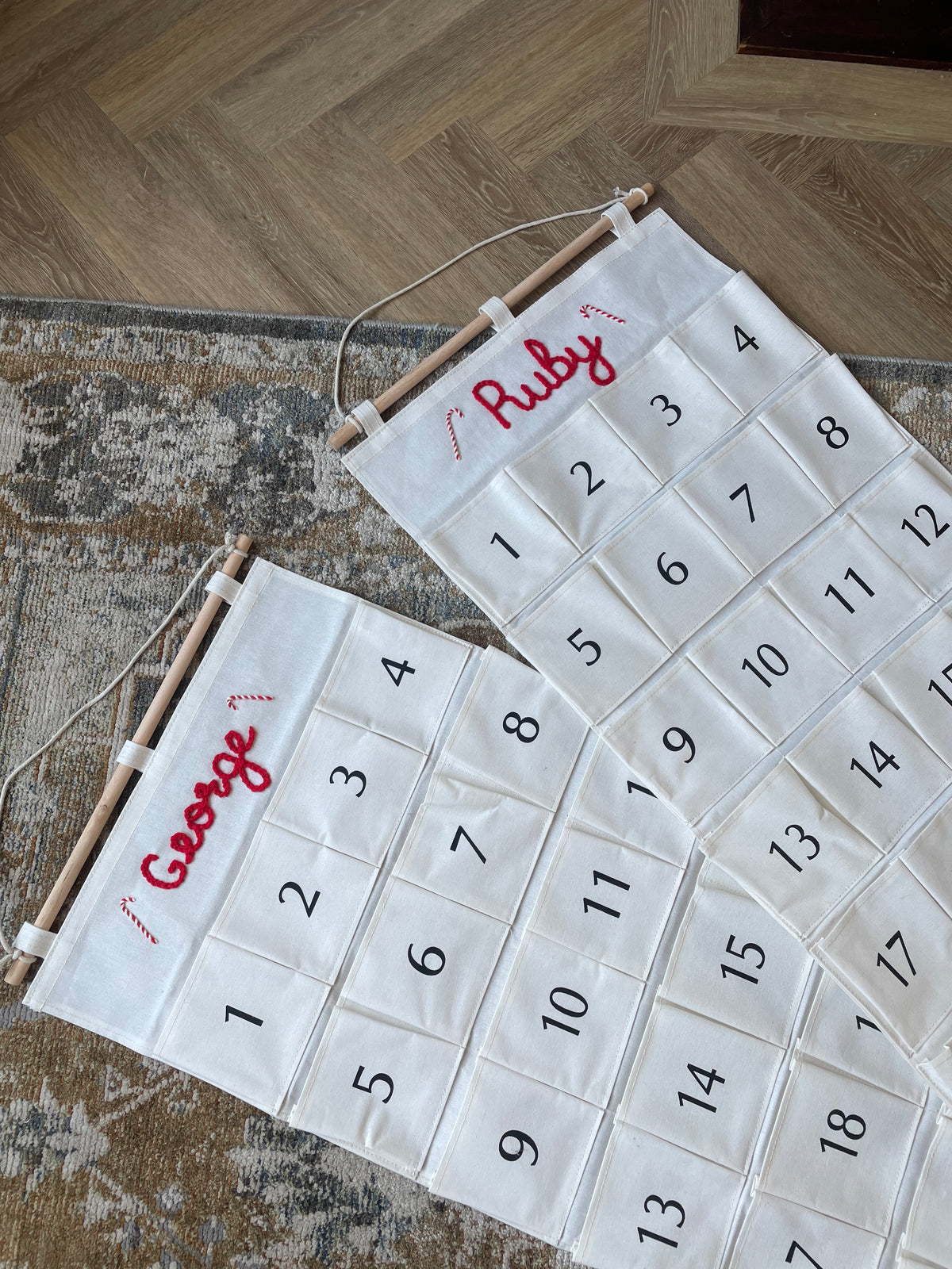 Two personalized Christmas advent calendars laid out on a patterned rug over a wood floor. One calendar is white with the name 'George' in red script and the other is white with the name 'Ruby' in red script, both with numbered pockets and wooden dowels.