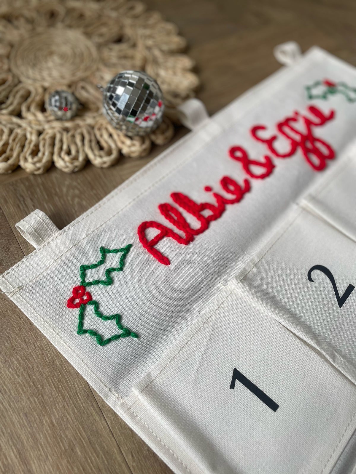 Two personalized Christmas advent calendars laid out on a patterned rug over a wood floor. One calendar is white with the name 'George' in red script and the other is white with the name 'Ruby' in red script, both with numbered pockets and wooden dowels