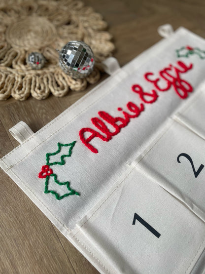 Two personalized Christmas advent calendars laid out on a patterned rug over a wood floor. One calendar is white with the name 'George' in red script and the other is white with the name 'Ruby' in red script, both with numbered pockets and wooden dowels